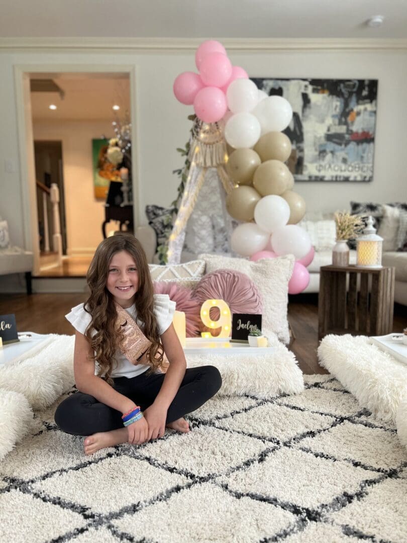 A girl sitting on a rug in a living room with balloons.